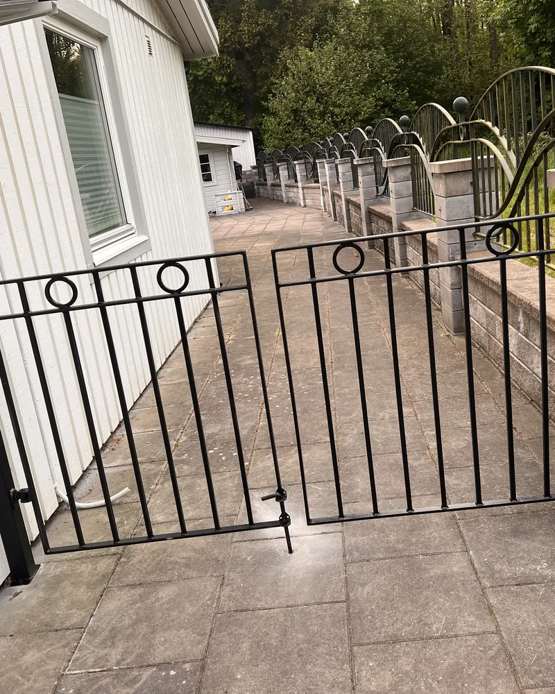 Svart dubbelgrind i smide med ringdekor och kulprydda stolpar, med bågformade smidesräcken längs stödmuren
