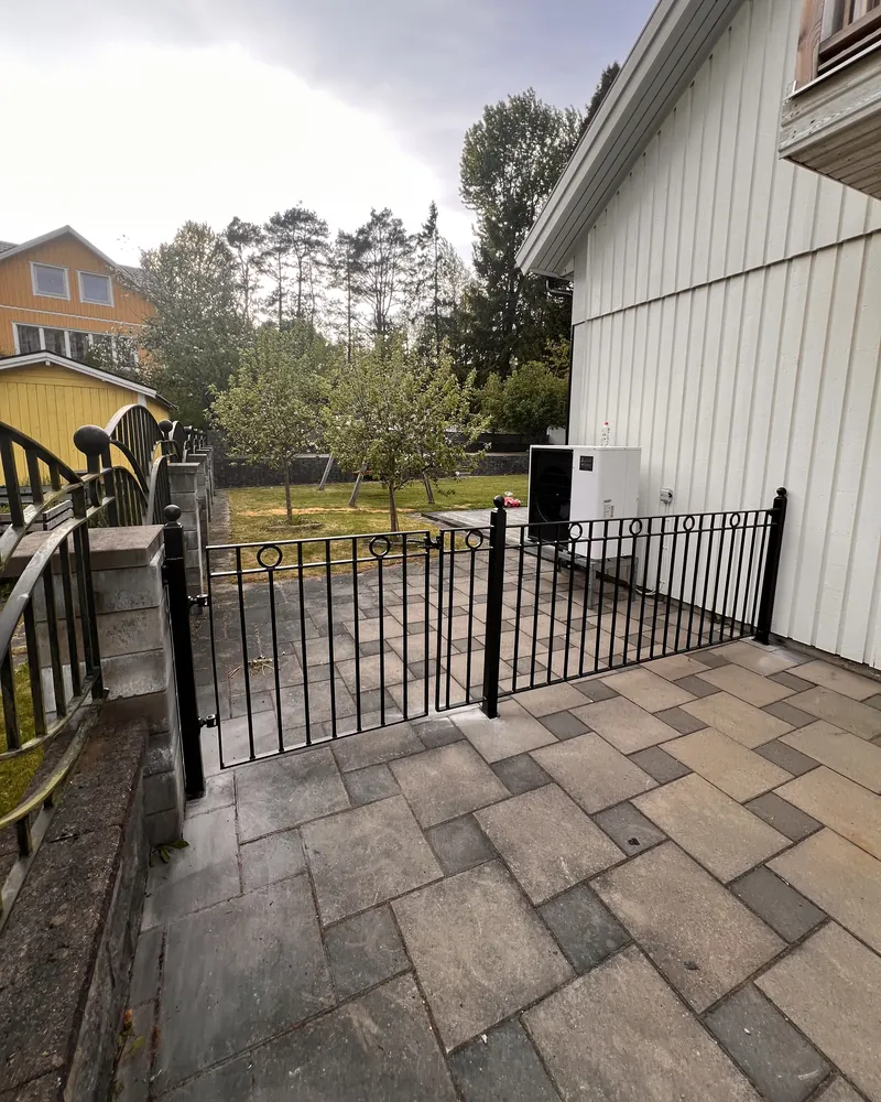 Svart smidesgrind och lågt staket med ringdekor över en stenlagd uteplats